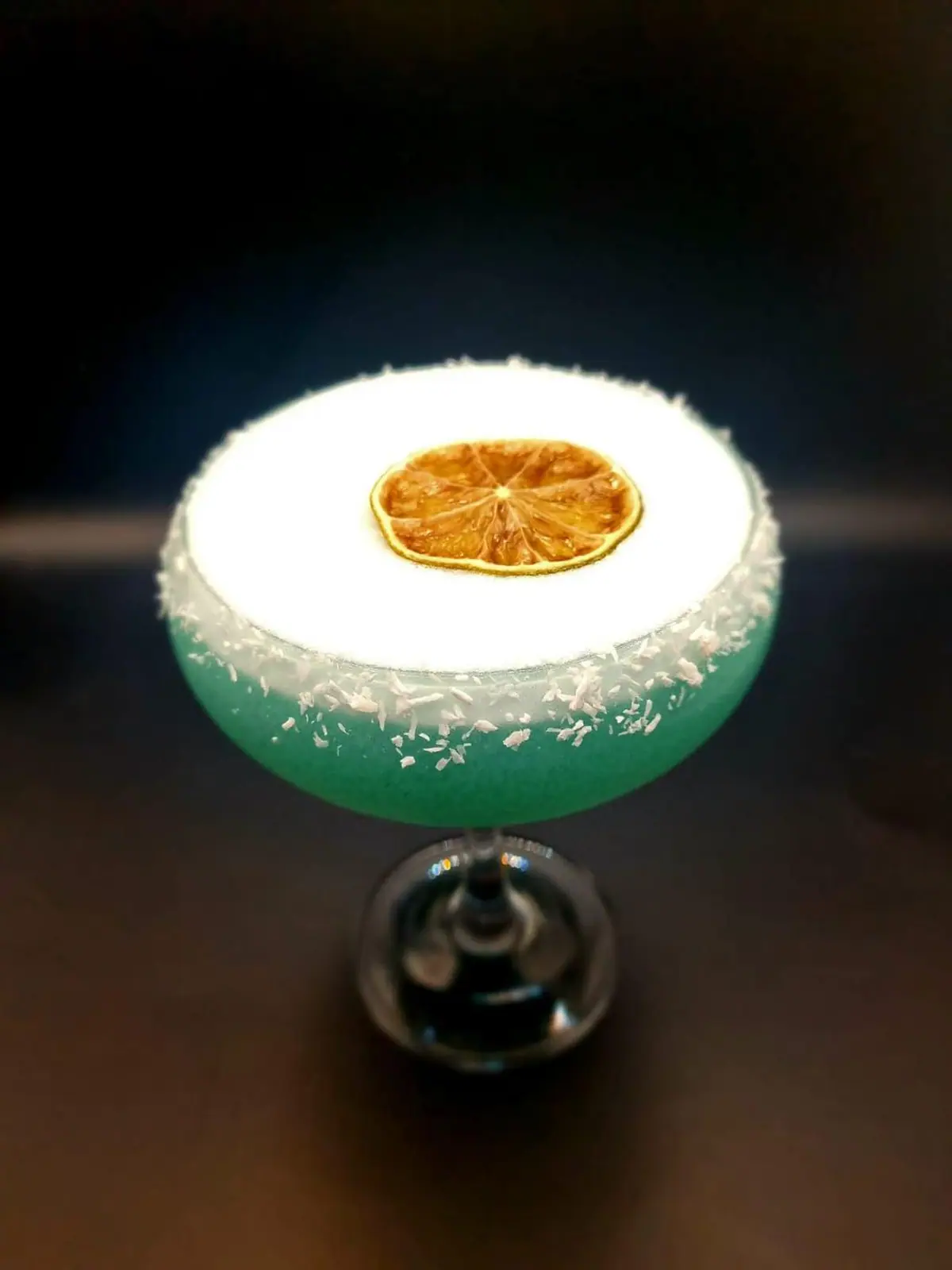A blue coupe cocktail with a coconut rim and dried orange