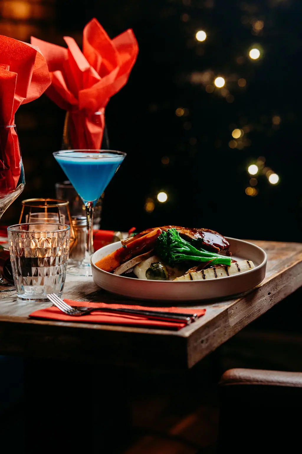 A teal cocktail and a festive plate on a candlelit table with Christmas lights